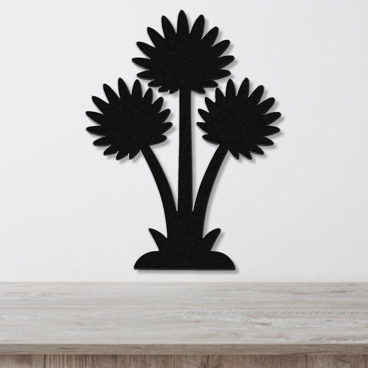 Black metal wall art cutout of three sunflower-like blooms with radiating petals, mounted on a white wall above a light wood surface.