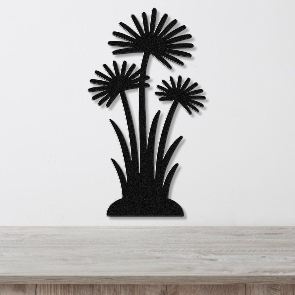 Black metal wall art featuring a silhouette of three daisy-like flowers with long stems and grassy base, mounted on a white wall above a wooden surface.