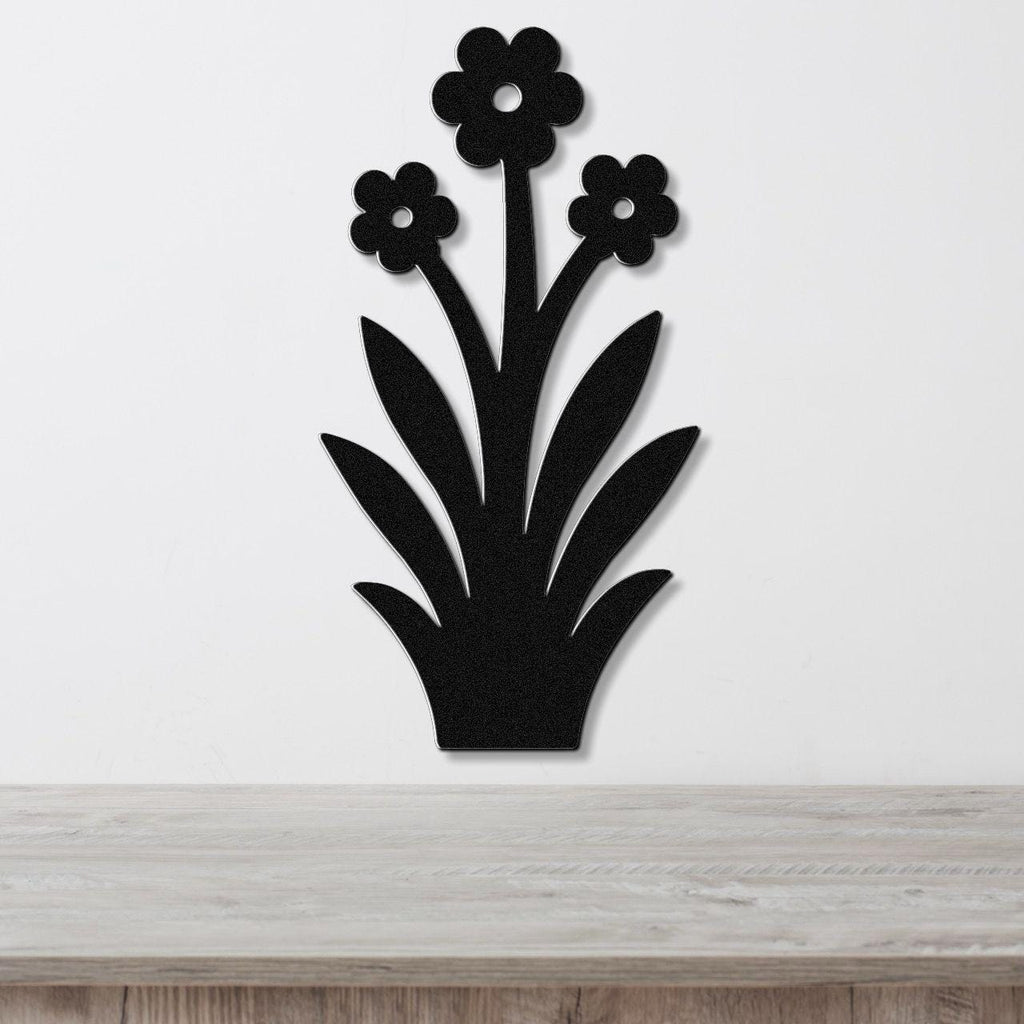 Black metal wall art silhouette of a wildflower with three blossoms and long leaves, mounted on a white wall above a wooden surface.