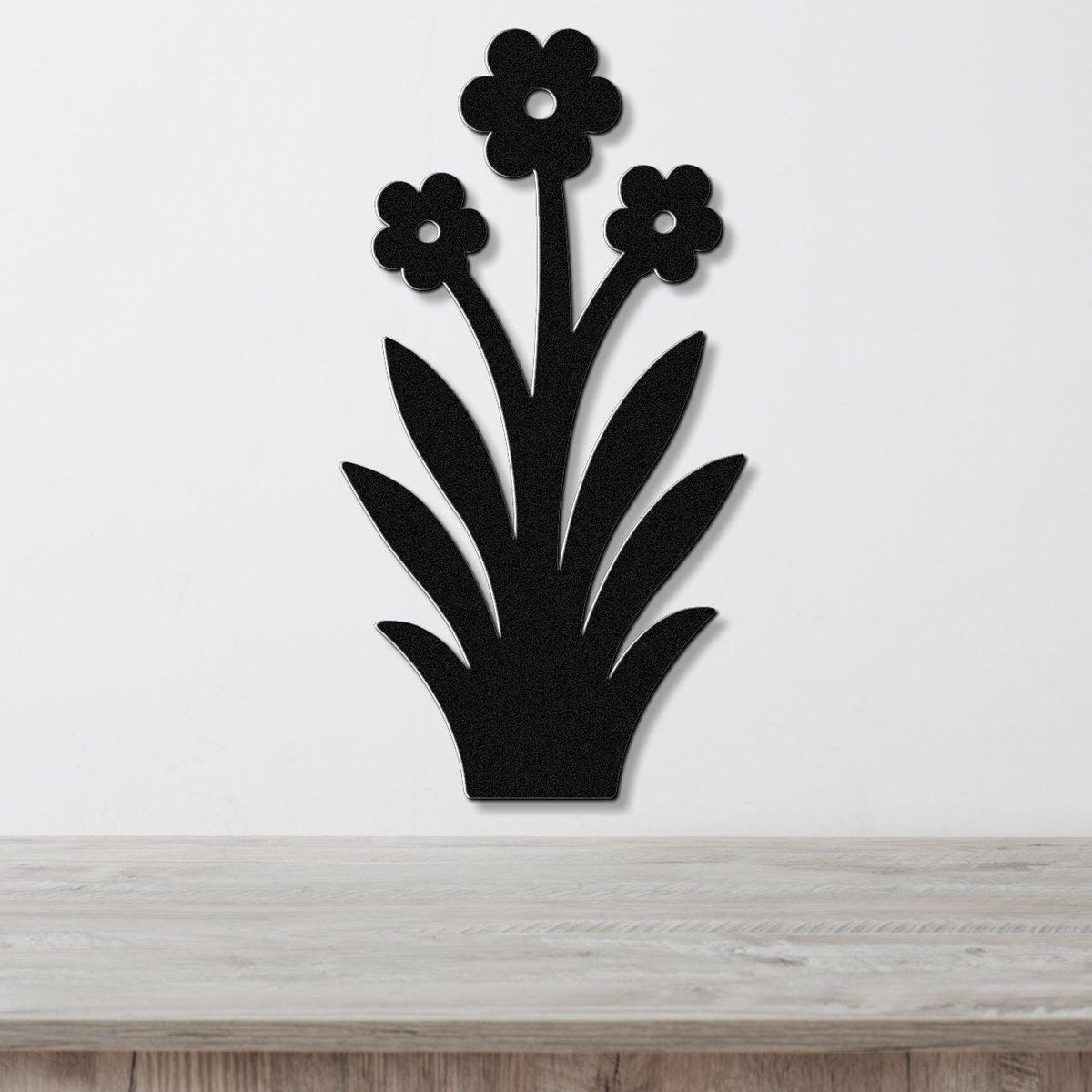 Black metal wall art silhouette of a wildflower with three blossoms and long leaves, mounted on a white wall above a wooden surface.