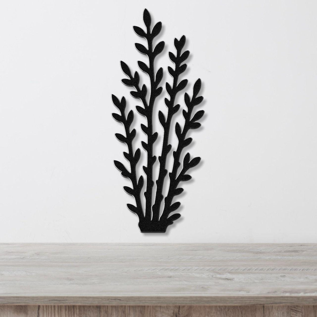 Black metal wall art in a silhouette of desert branches with layered leaf cutouts, mounted on a white wall above a light wood surface.