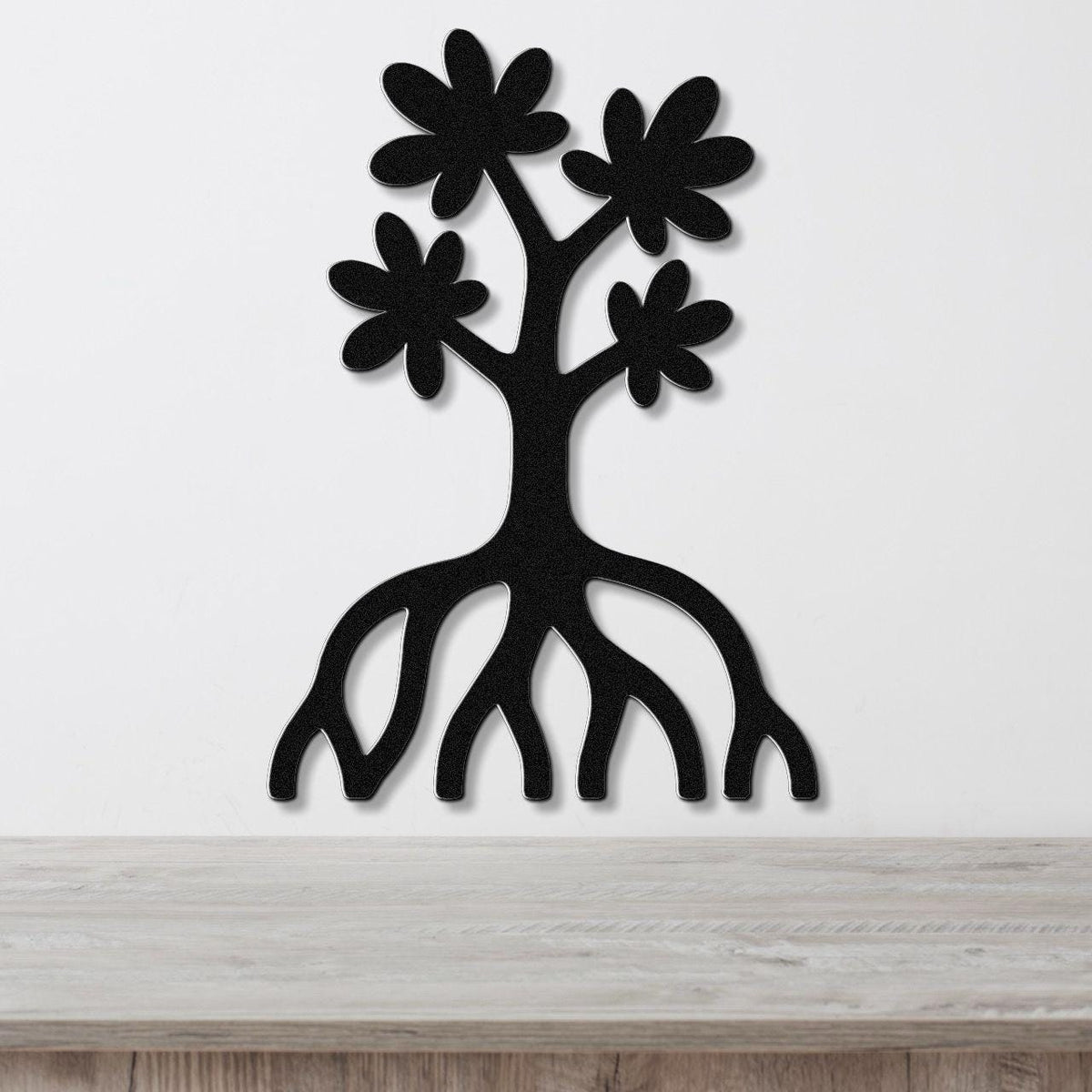 Black metal wall art in the shape of a tree with flower cutouts and exposed roots, mounted on a white wall above a wooden surface.