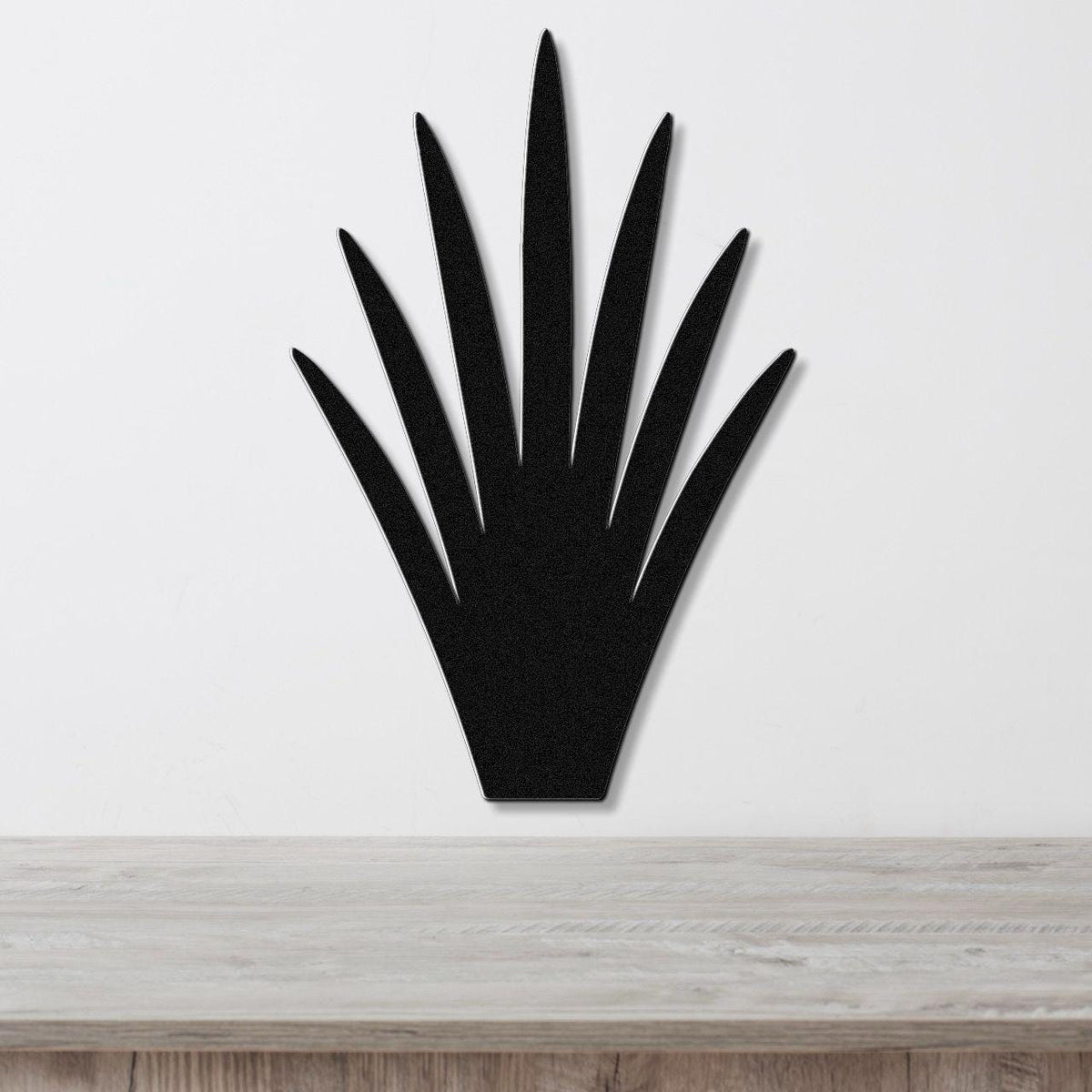 Black metal yucca wall art with pointed leaf cutouts, mounted on a white wall above a light wood surface.