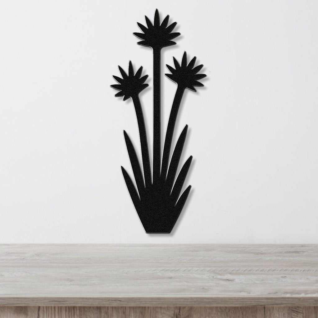 Black metal wall art in a yucca plant silhouette with three starburst flowers and spiky leaves, mounted on a white wall above a light wood surface.