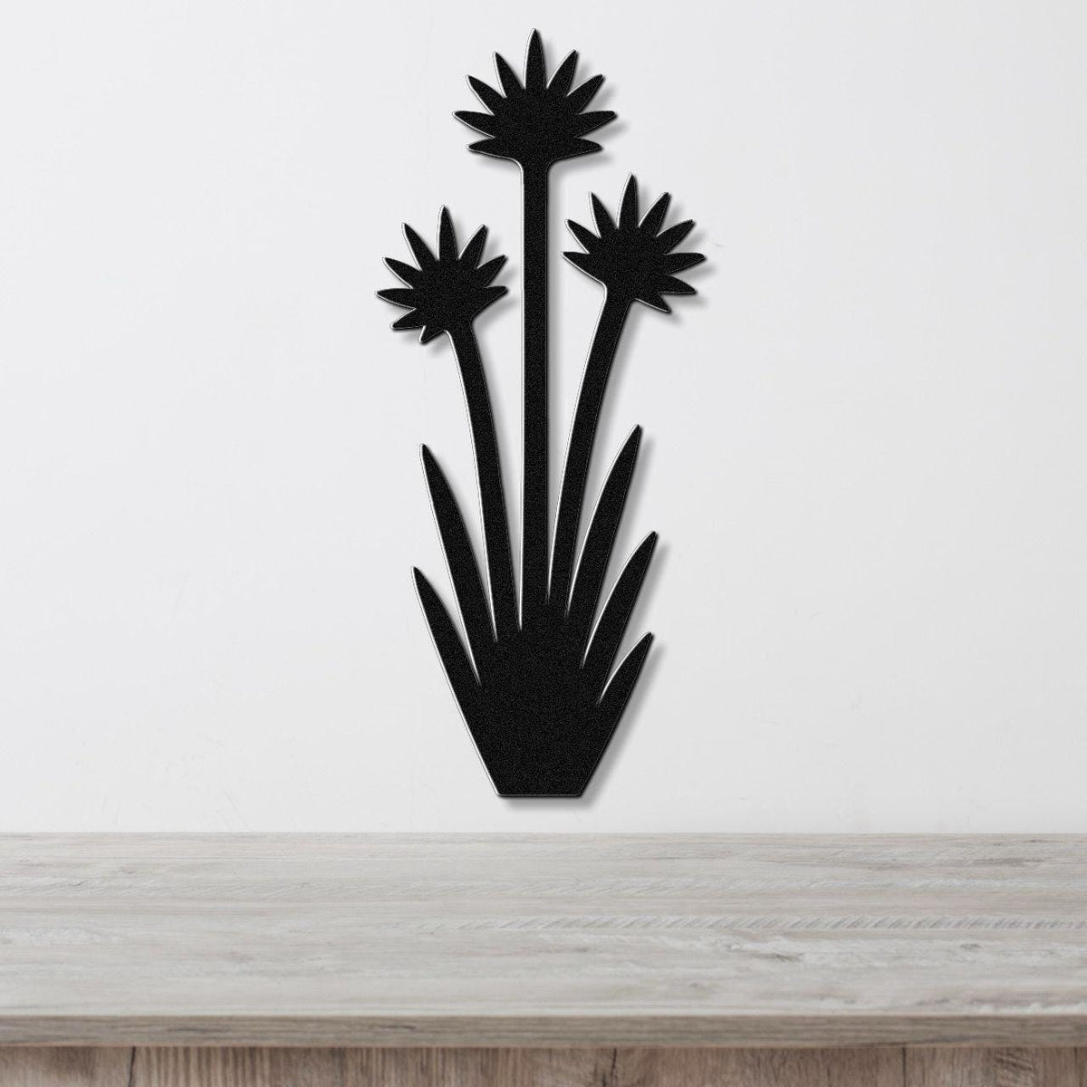Black metal wall art in a yucca plant silhouette with three starburst flowers and spiky leaves, mounted on a white wall above a light wood surface.
