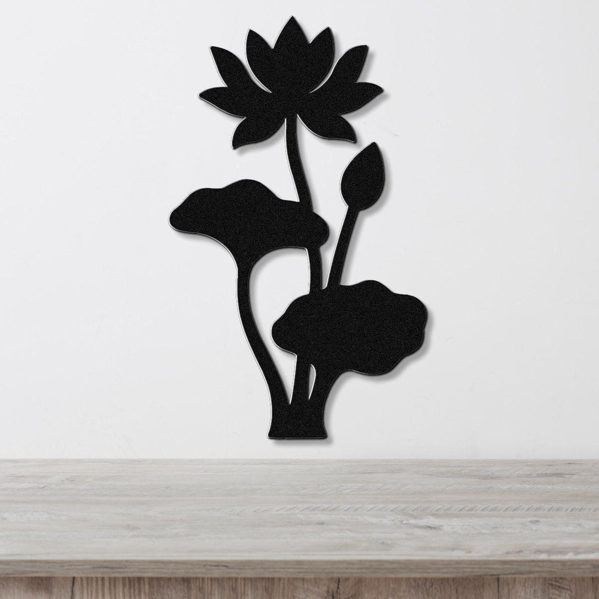 Black metal lotus wall art with a flower, bud, and leaves in a silhouette cutout style, mounted on a white wall above a wooden surface.
