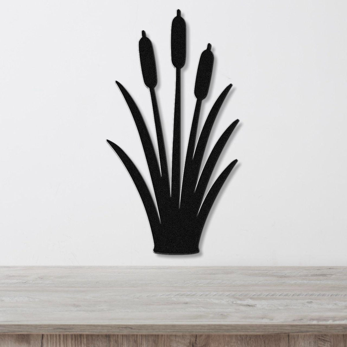 Black metal cattail wall art silhouette with three seed heads and long leaves, mounted on a white wall above a light wood surface.