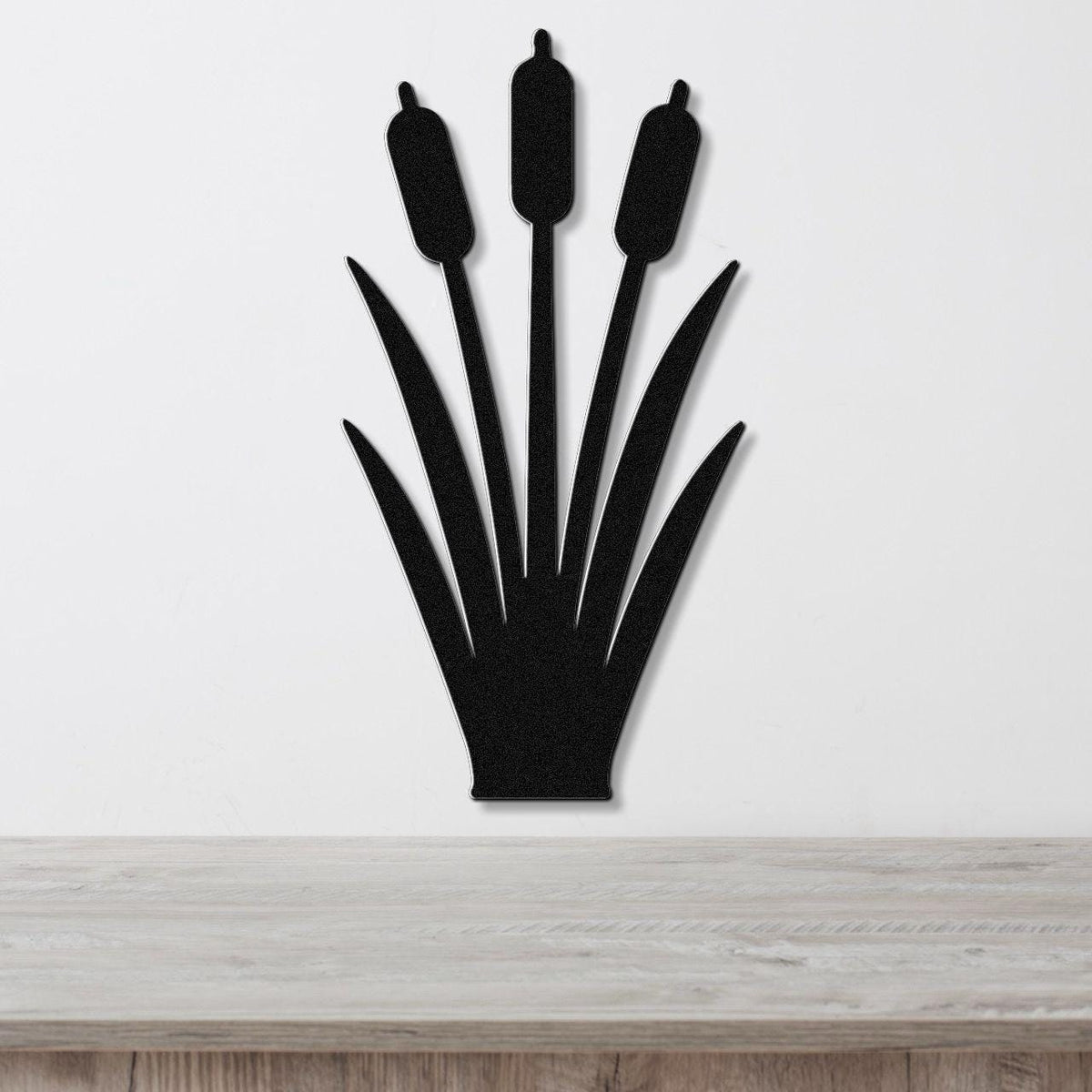 Black metal cattail wall art silhouette with three seed heads and long leaves, mounted on a white wall above a light wood surface.