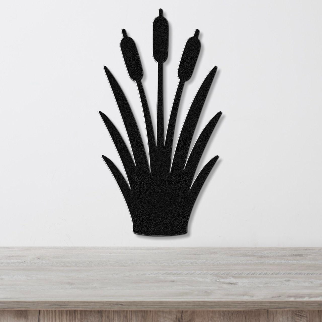 Black metal cattail wall art with a silhouette design of three seed heads and long leaves, mounted on a white wall above a light wood surface.