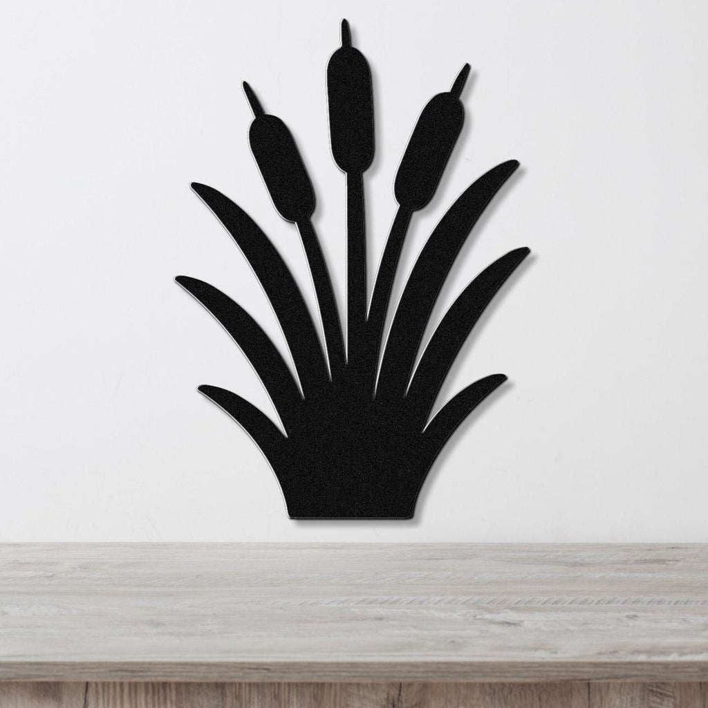 Black metal cattail wall art silhouette with three seed heads and long leaves, mounted on a white wall above a light wood surface.