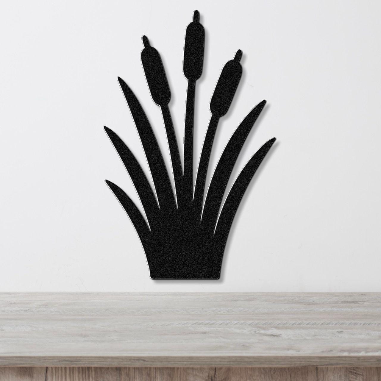 Black metal cattail wall art with a silhouette design of three seed heads and long leaves, mounted on a white wall above a light wood surface.