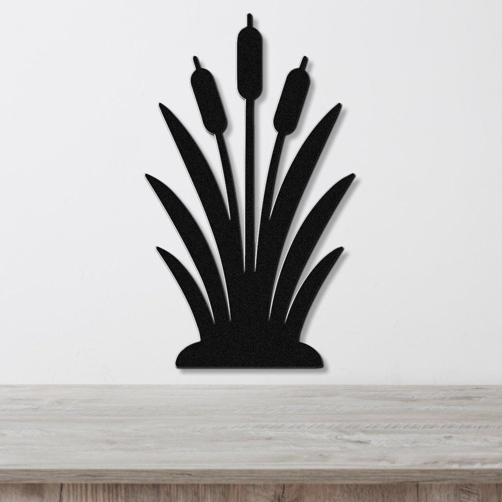 Black metal wall art in the silhouette of a cattail plant with three seed heads and long leaves, mounted on a white wall above a wooden surface.