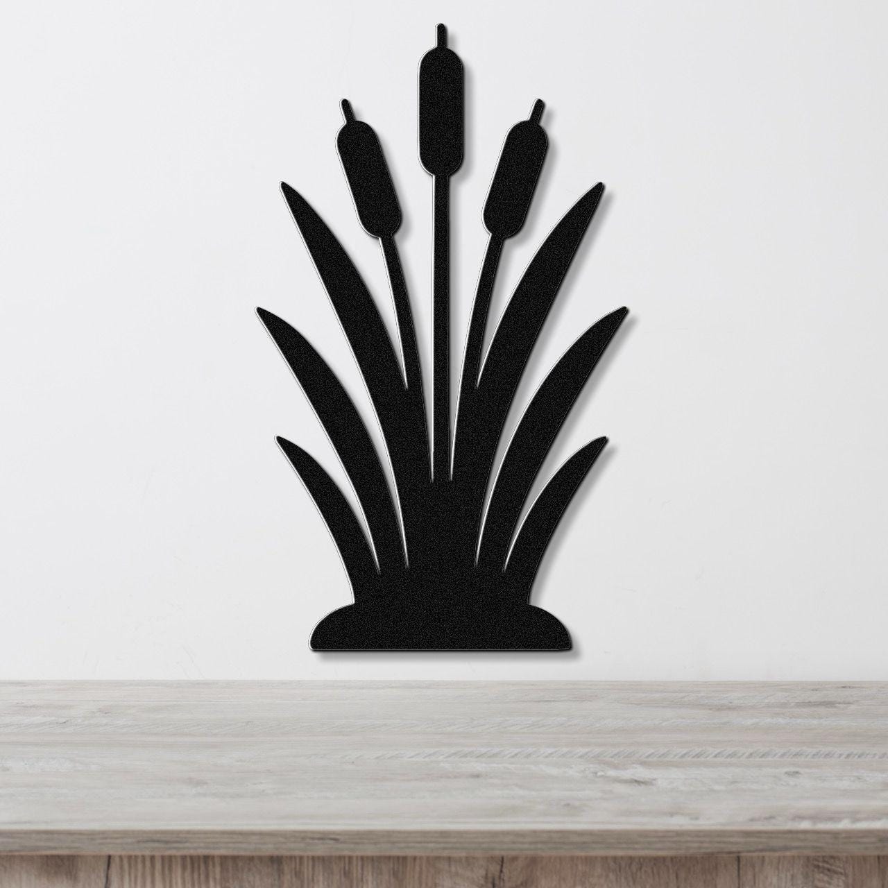 Black metal wall art in the silhouette of a cattail plant with three seed heads and long leaves, mounted on a white wall above a wooden surface.
