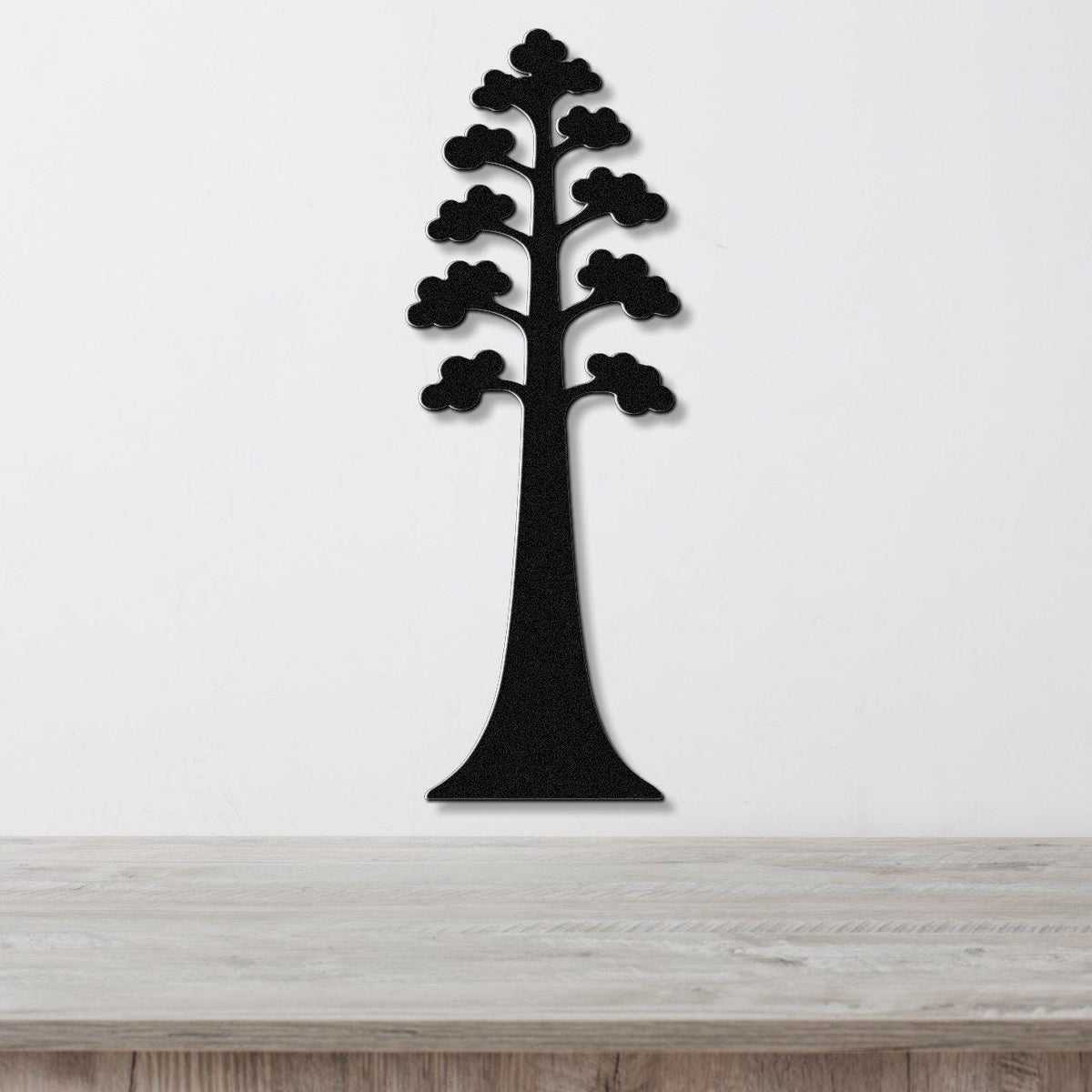 Black metal wall art in the silhouette of a sequoia tree with layered branches, mounted on a white wall above a wooden surface.