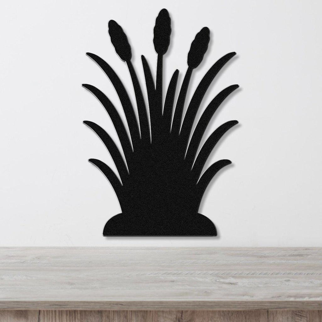 Black metal wall art cutout of wild grass with three seed heads, mounted on a white wall above a light wood surface.