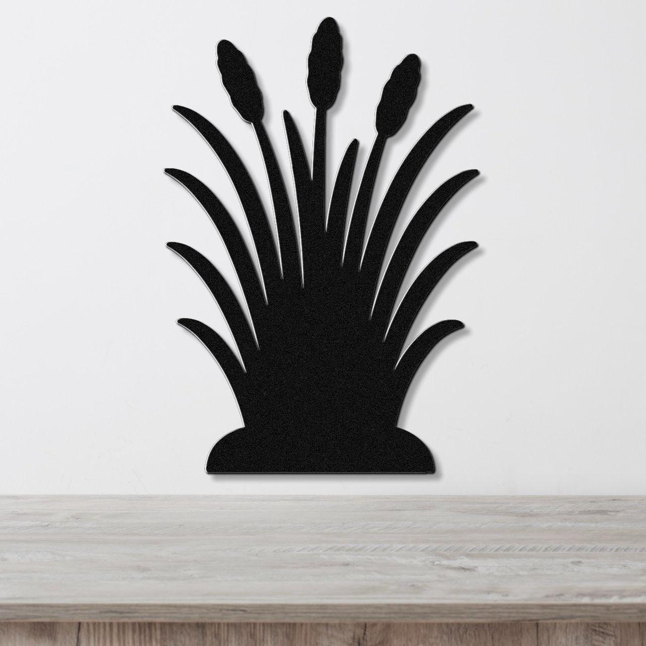 Black metal wall art cutout of wild grass with three seed heads, mounted on a white wall above a light wood surface.