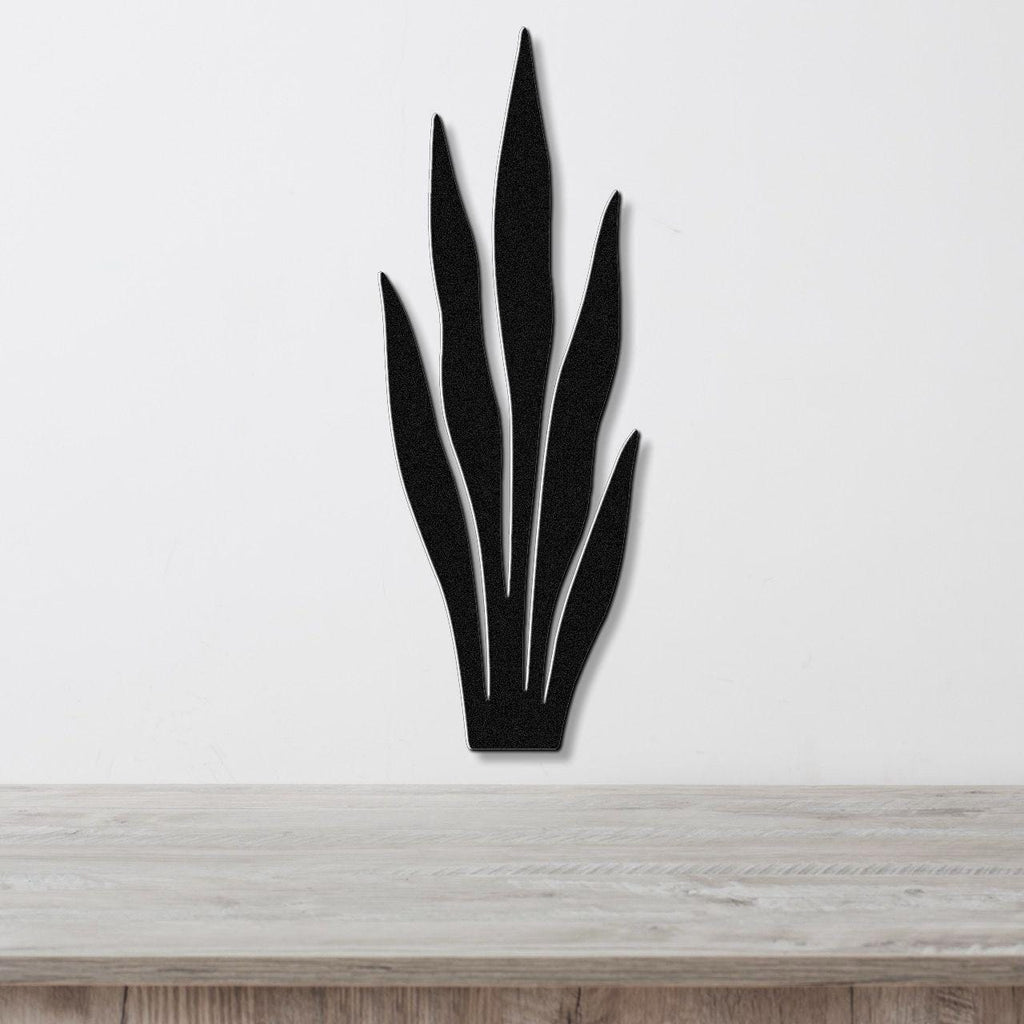 Black metal wall art in the shape of a snake plant with internal cutout lines, mounted on a white wall above a light wood surface.