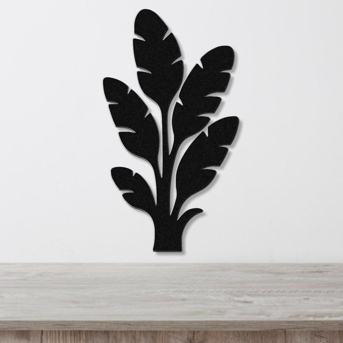 Black metal wall art in a tropical leaf silhouette design with layered cutouts, mounted on a white wall above a light wood surface.