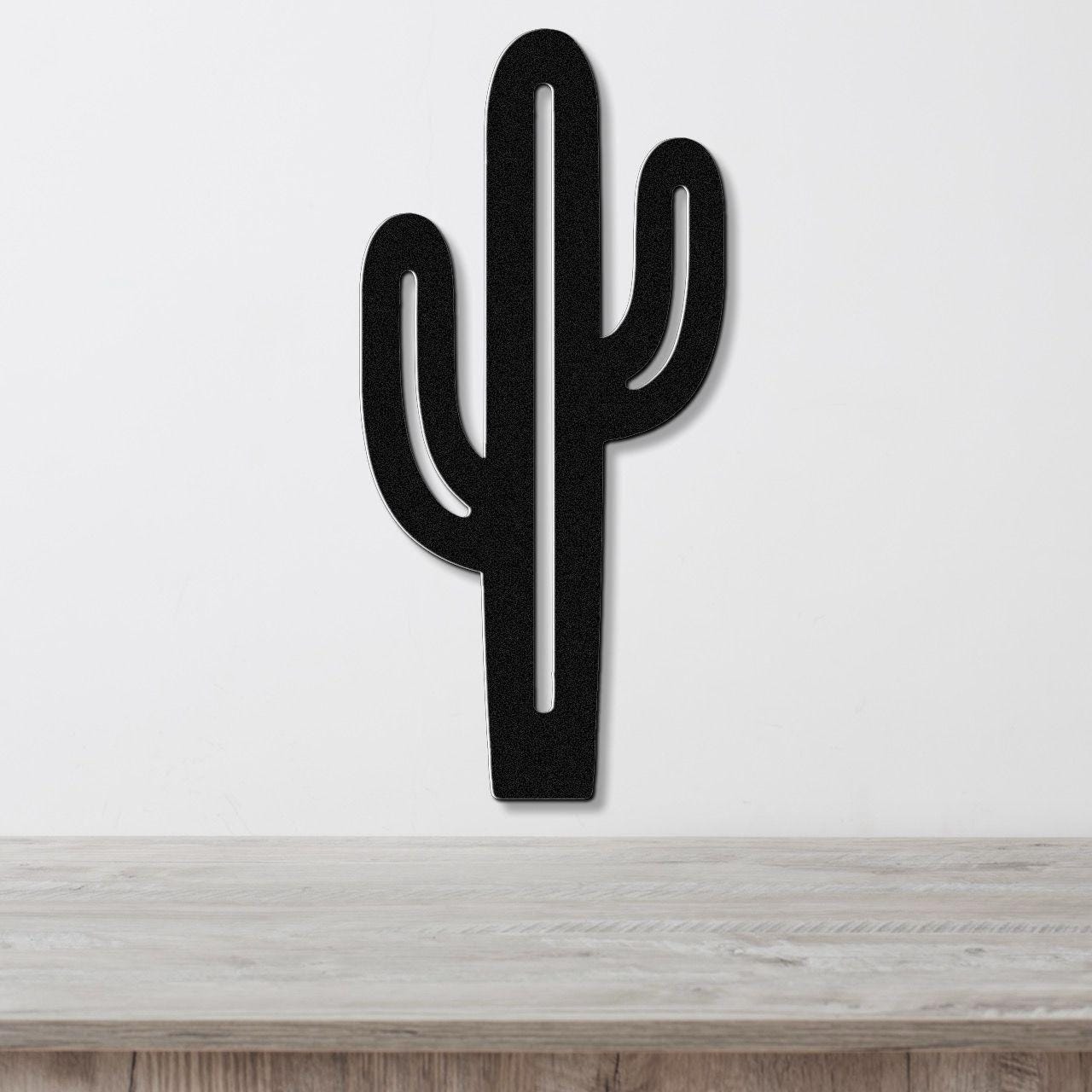 Black metal saguaro cactus wall art with cutout arms, mounted on a white wall above a light wood surface.