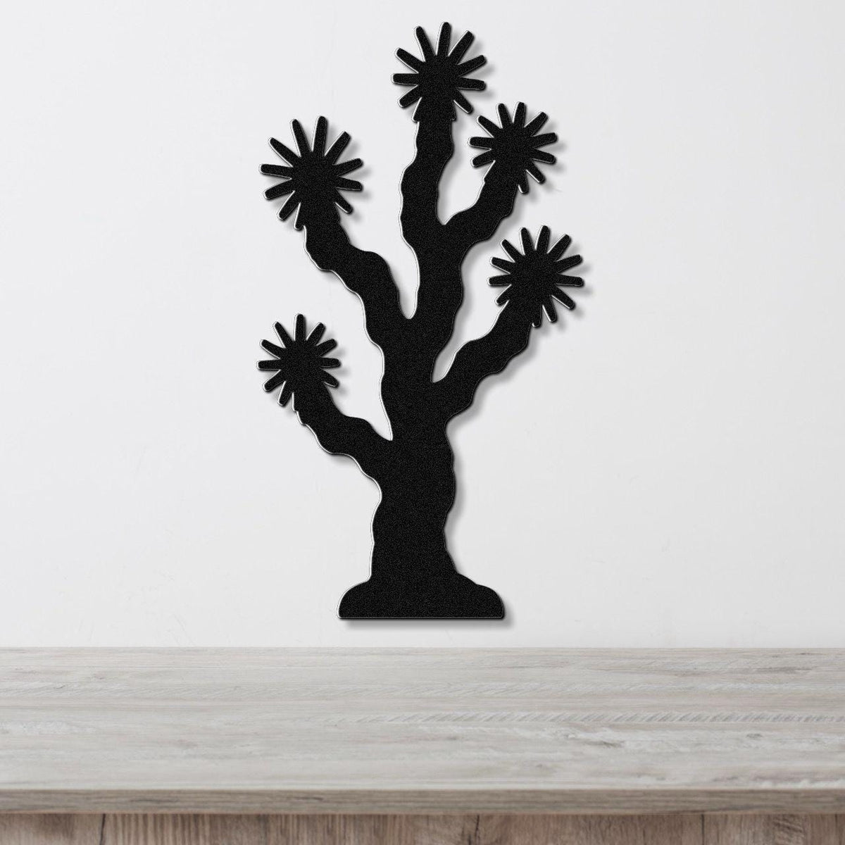 Black metal wall art cutout of a Joshua tree with starburst-shaped branches, mounted on a white wall above a light wood surface.