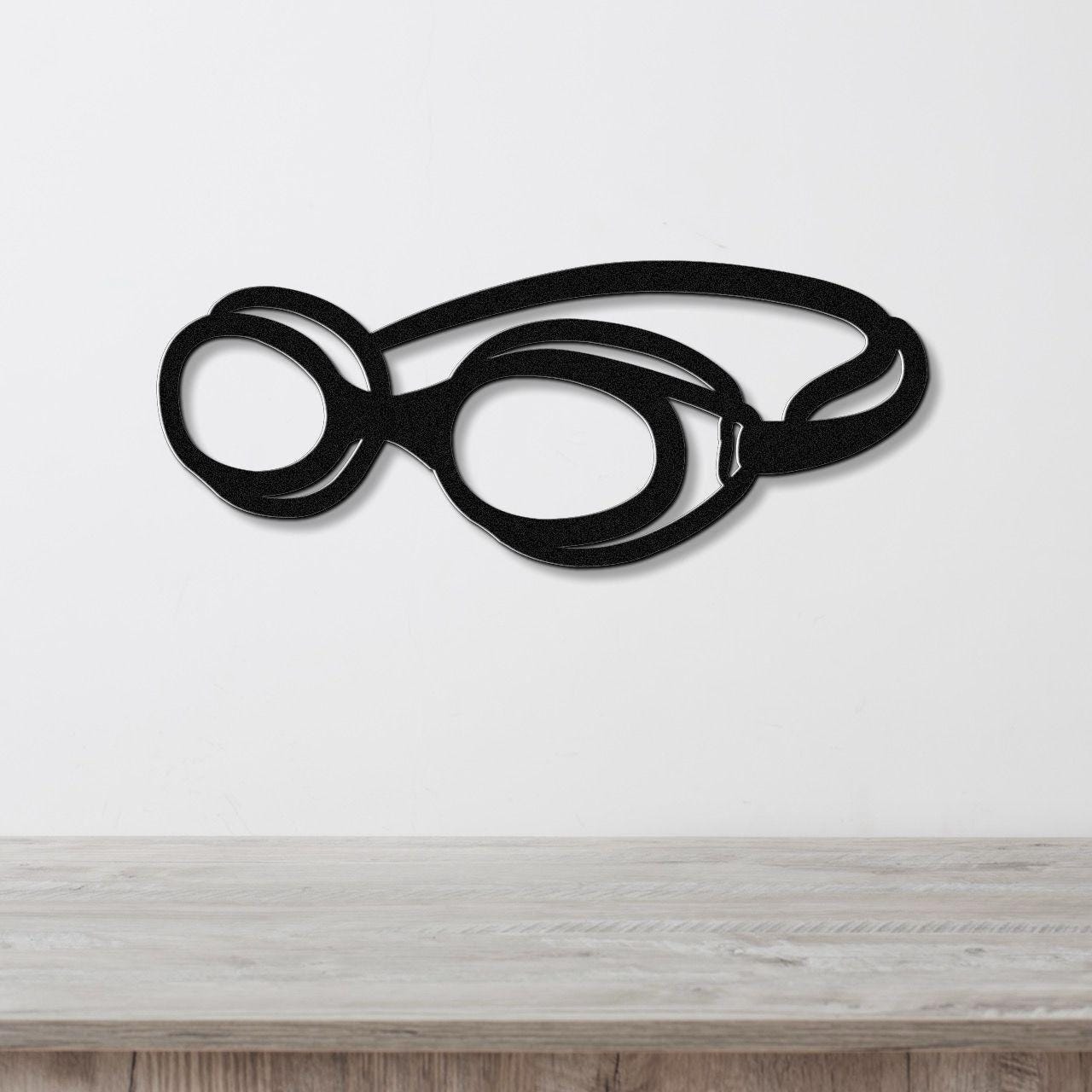 Black metal wall art shaped like swim goggles with cutout lenses and straps, mounted on a white wall above a light wood surface.