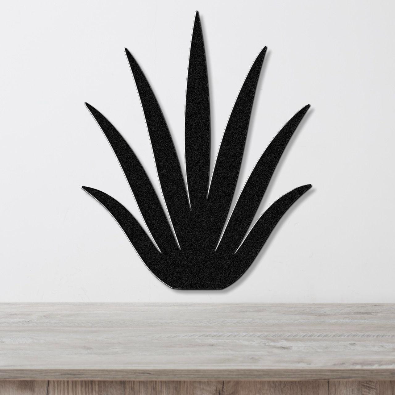 Black metal wall art cutout of a century plant with spiky leaves, mounted on a white wall above a light wood surface.
