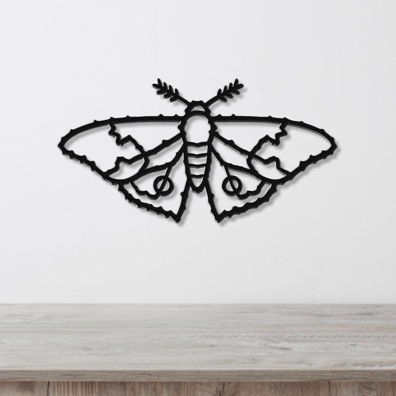 Black metal moth wall art with detailed wing cutouts and leafy antennae, mounted on a white wall above a wooden surface.