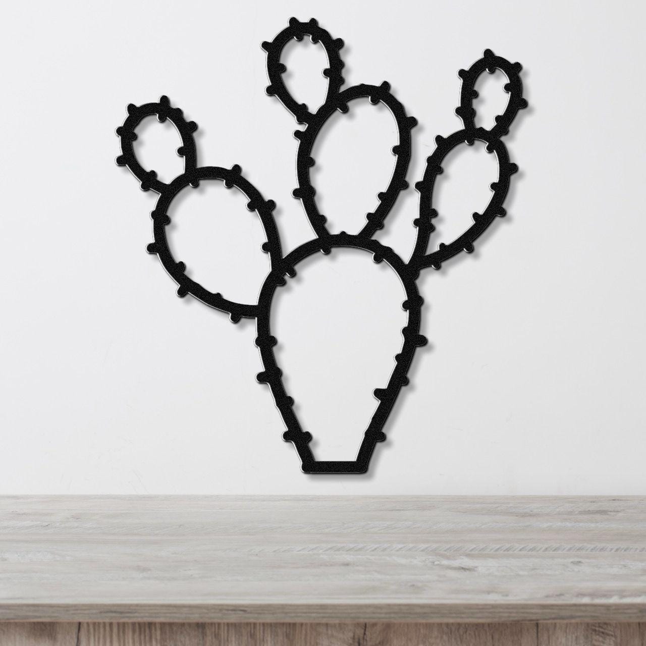 Black metal wall art cutout of a prickly pear cactus with spiny edges, mounted on a white wall above a light wood surface.