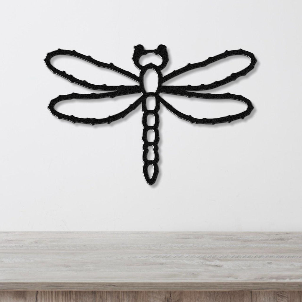 Black metal dragonfly wall art with cutout wings and segmented body, mounted on a white wall above a light wood surface.