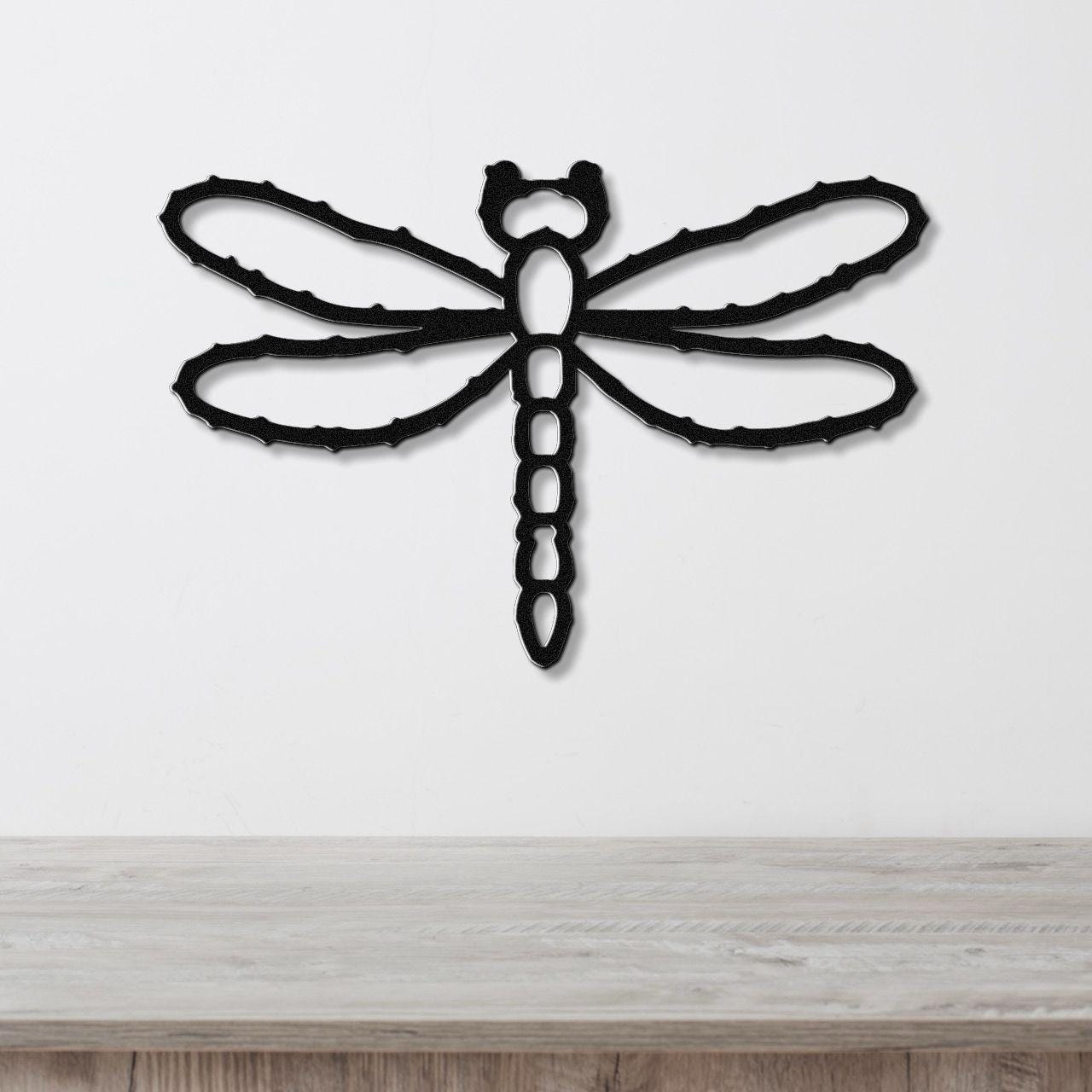 Black metal dragonfly wall art with cutout wings and segmented body, mounted on a white wall above a light wood surface.