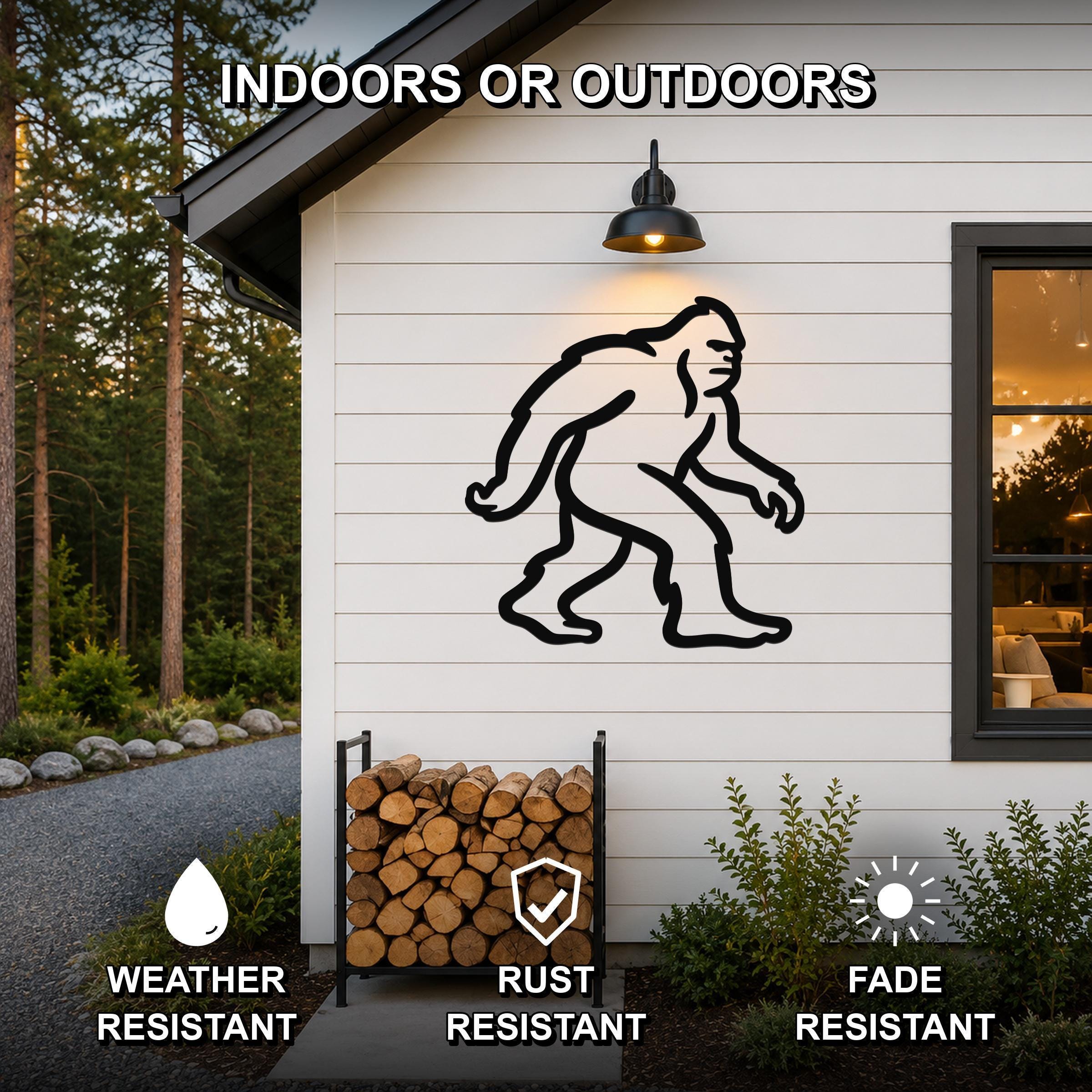 Black metal Bigfoot silhouette wall art mounted on a white exterior wall above a firewood rack, with text indicating indoor or outdoor use and weather, rust, and fade resistance.