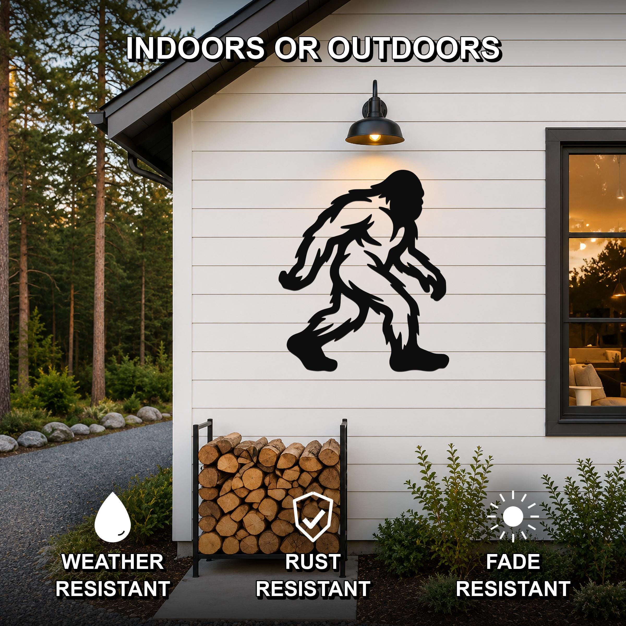 Black metal Bigfoot silhouette wall art mounted on a white siding exterior wall above a firewood rack, with text indicating it is weather, rust, and fade resistant for indoor or outdoor use.