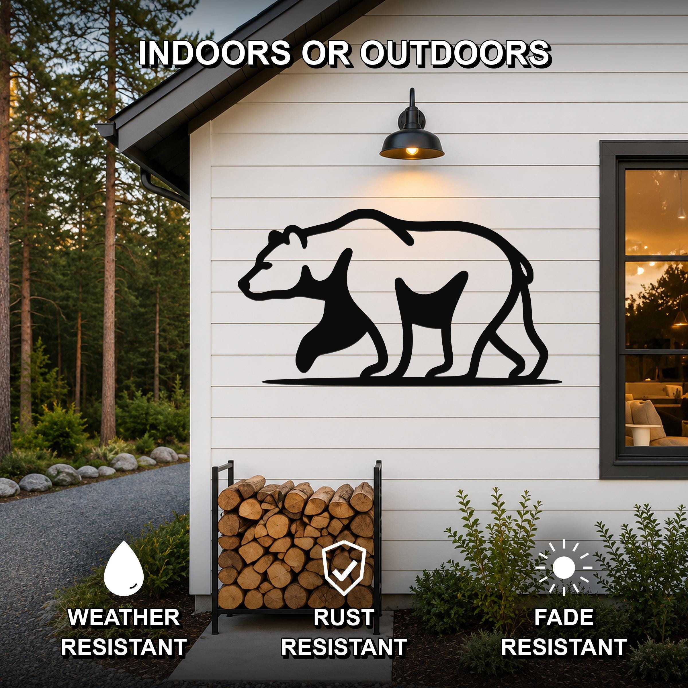 Black metal bear silhouette wall art mounted on a white siding exterior wall above a firewood rack, with text indicating it is weather, rust, and fade resistant for indoor or outdoor use.