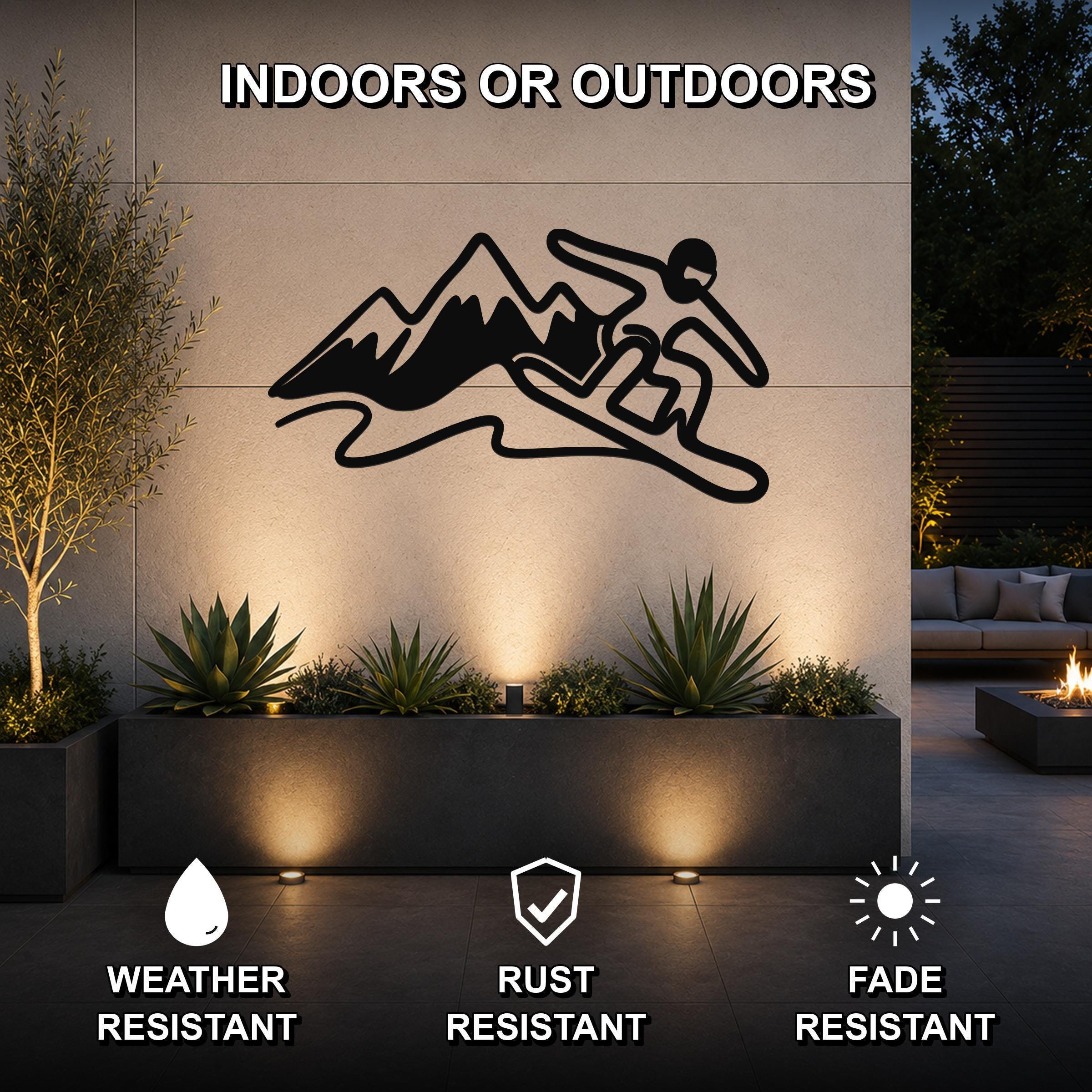 Black metal wall art silhouette of a snowboarder in motion above mountains, mounted on an outdoor wall above a planter with succulents and uplighting.