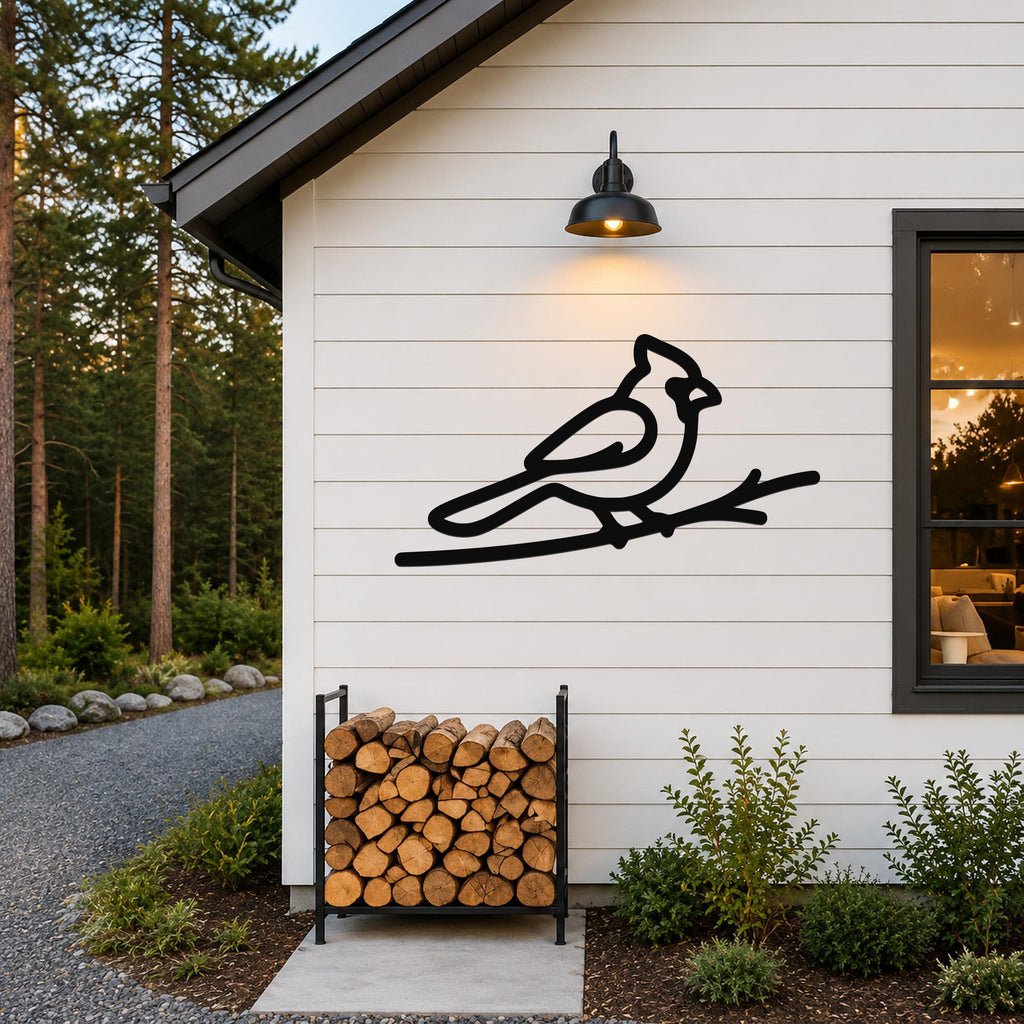 Black metal cardinal wall art in a bold silhouette, perched on a branch, mounted on a white exterior wall above a firewood rack.