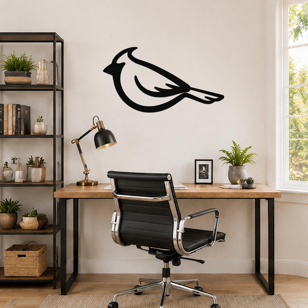 Black metal cardinal wall art silhouette mounted above a wooden desk in a home office, with a black office chair and a bookshelf nearby.
