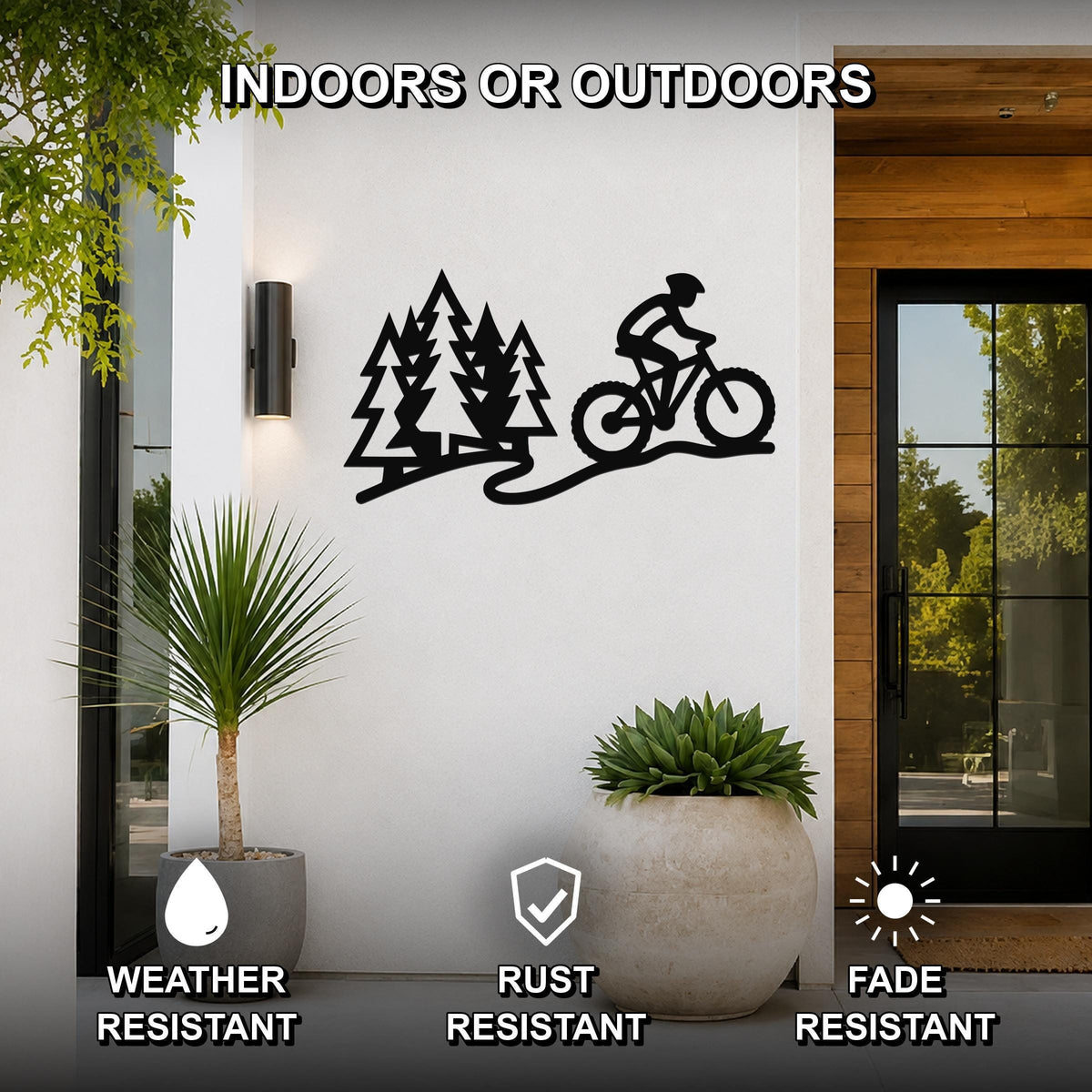 Black metal wall art featuring a cyclist and pine trees in a silhouette cutout style, mounted on a white exterior wall beside a glass door and potted plants.