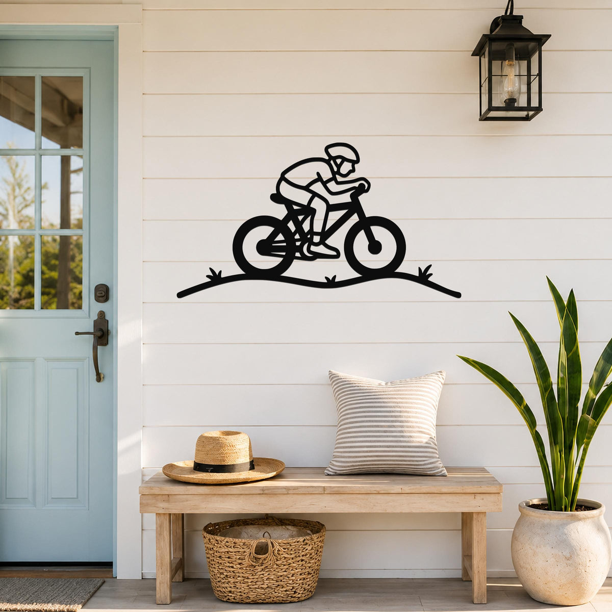 Black metal wall art silhouette of a cyclist on a trail, mounted on a white shiplap wall above a wooden bench in a porch entryway.
