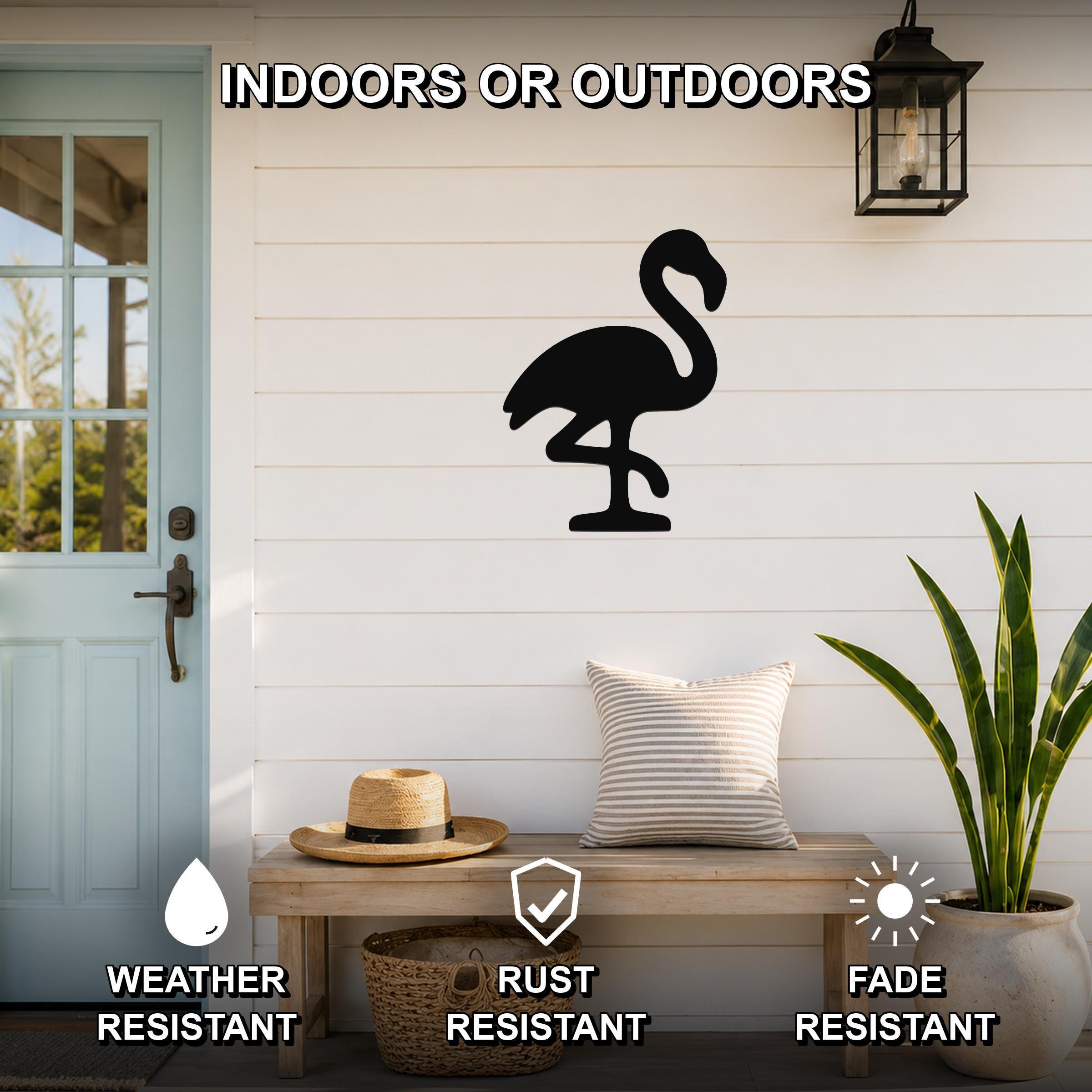 Black metal flamingo wall art silhouette mounted on a white shiplap wall above a wooden bench on a porch, with text indicating it is weather, rust, and fade resistant.
