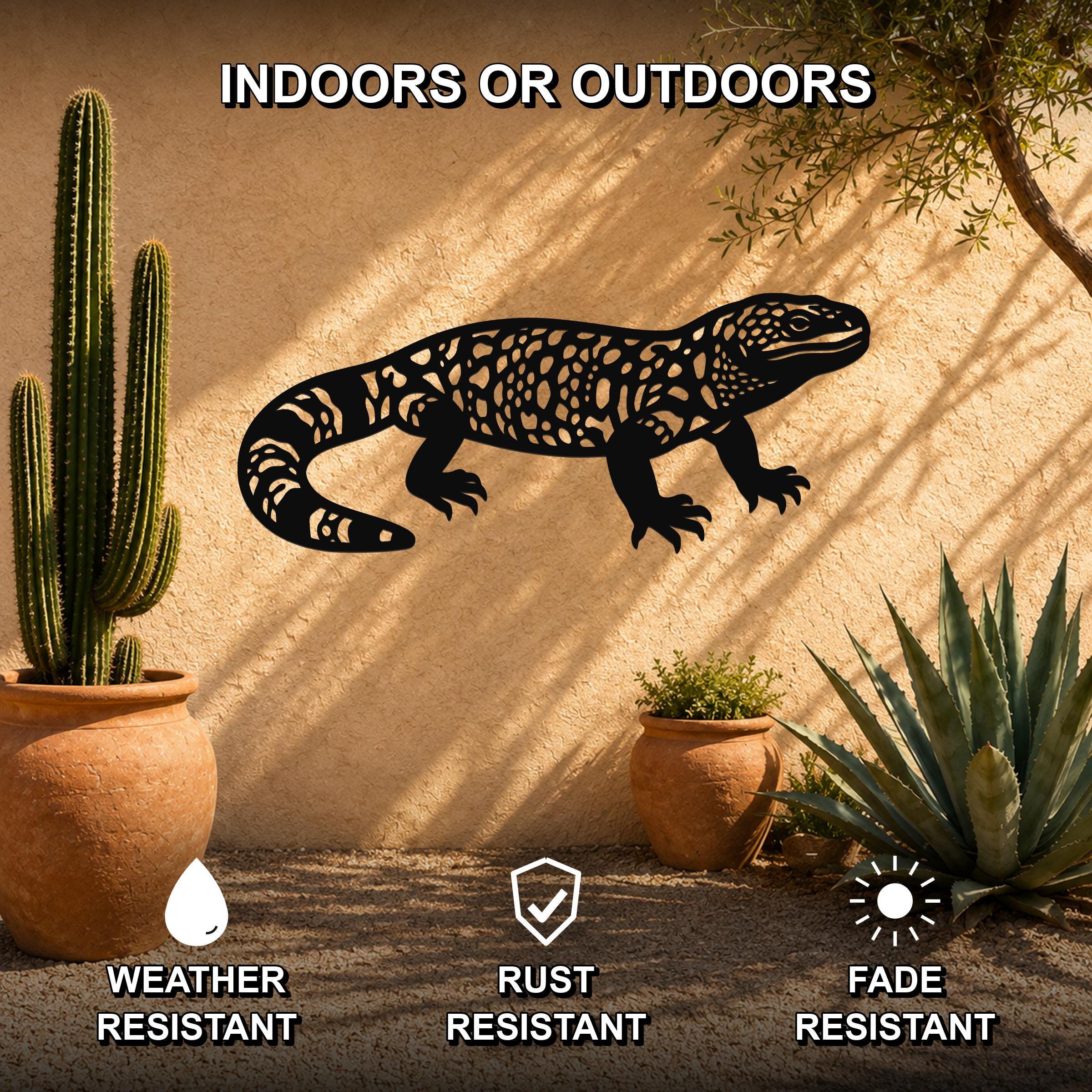 Black metal Gila monster wall art with intricate cutout patterns, mounted on a stucco outdoor wall beside potted cacti and succulents.