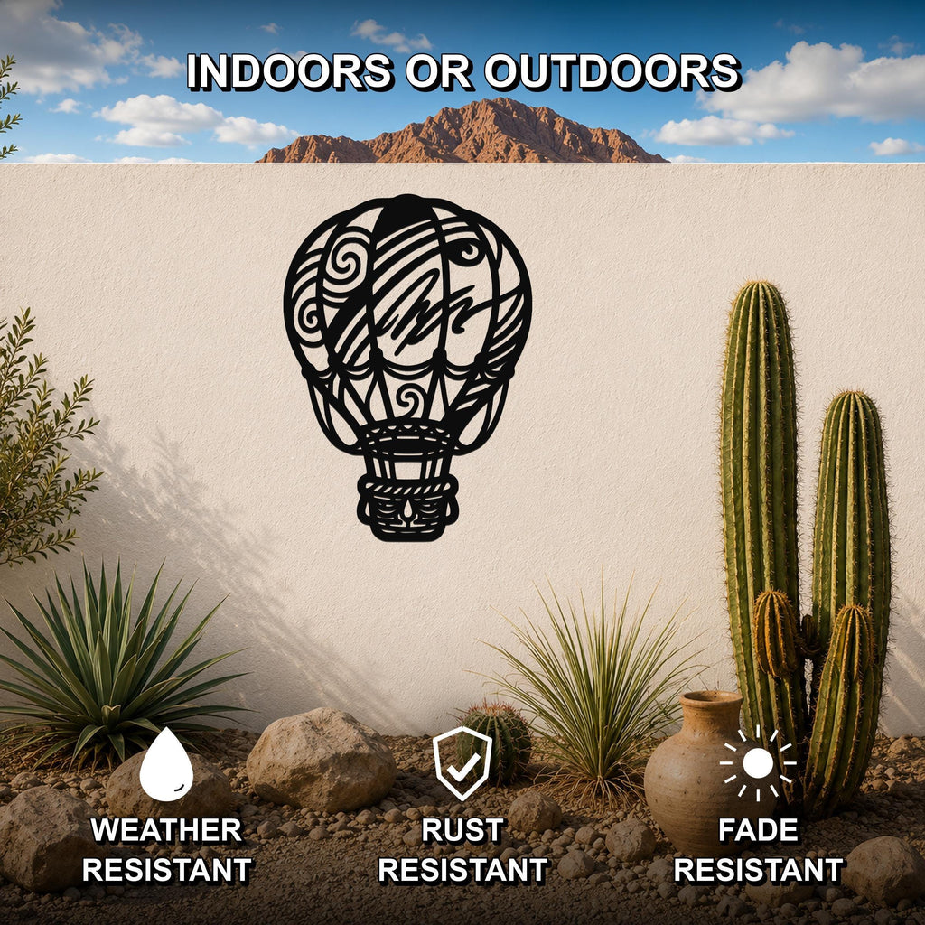 Black metal hot air balloon wall art with intricate swirl and line cutouts, mounted on a white outdoor wall above a desert garden with cacti and rocks.