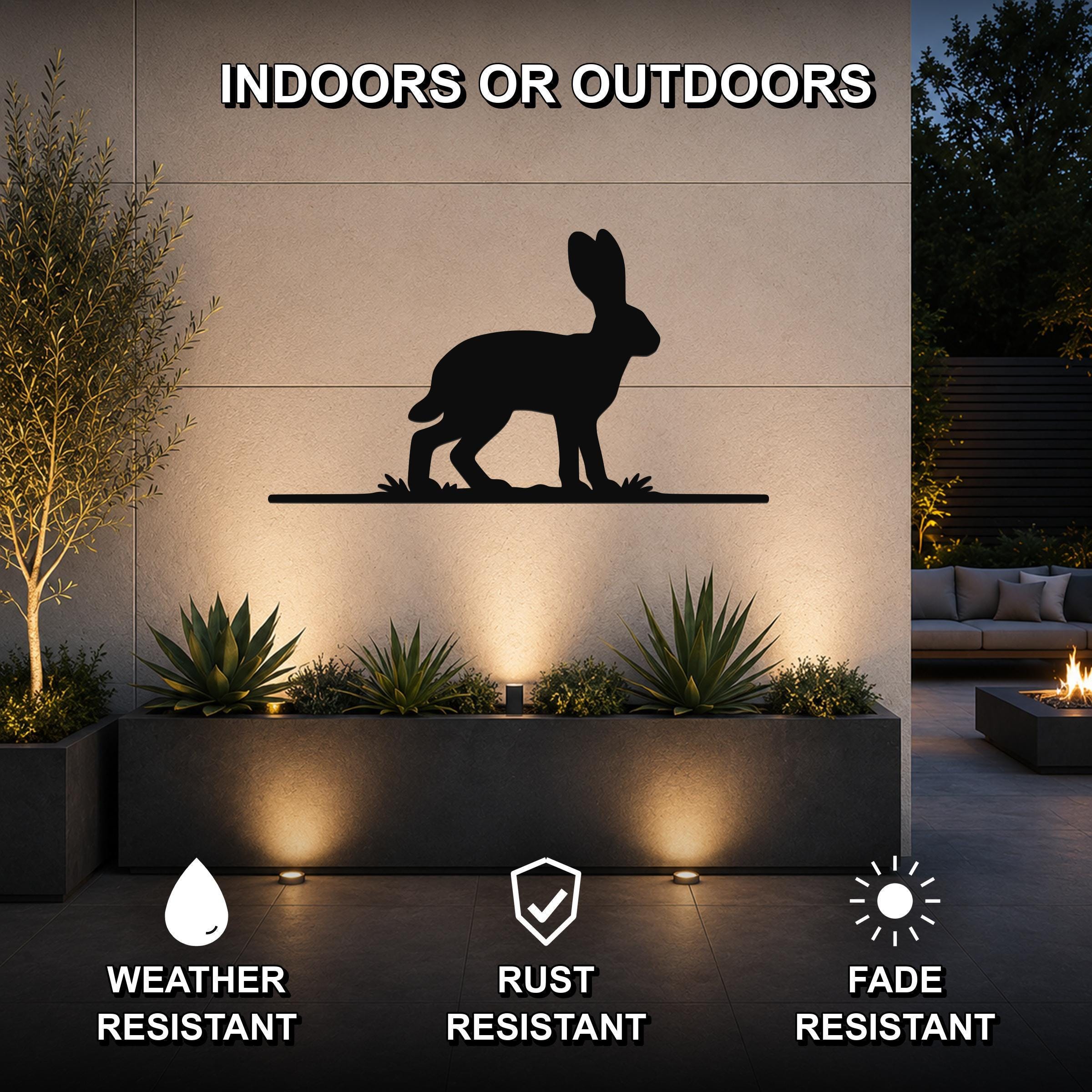 Black metal jackrabbit wall art silhouette mounted on an outdoor wall above a planter, with text indicating it is weather, rust, and fade resistant for indoor or outdoor use.