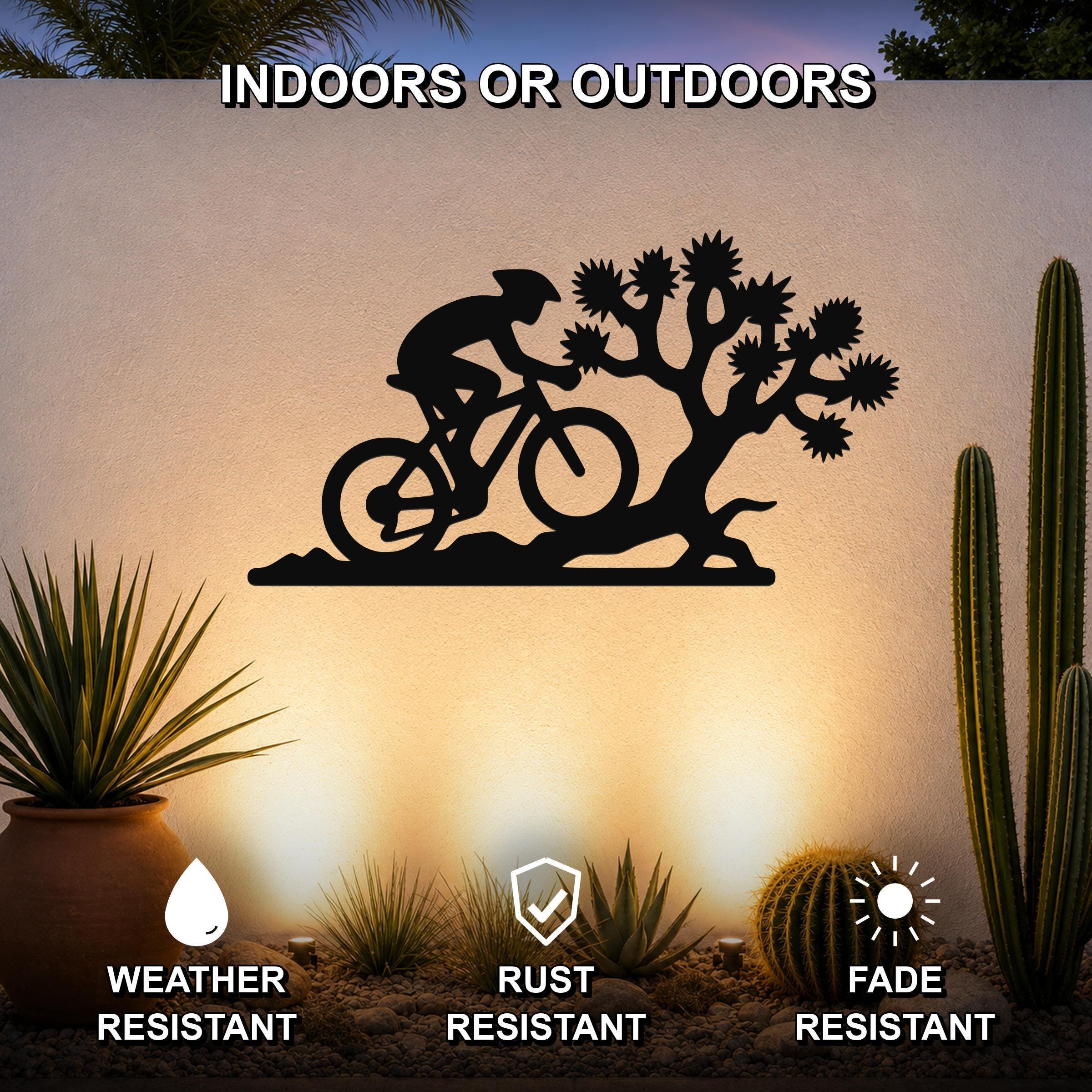 Black metal wall art featuring a cyclist silhouette and Joshua tree cutout, mounted on an outdoor wall above a desert garden with cacti and potted plants.