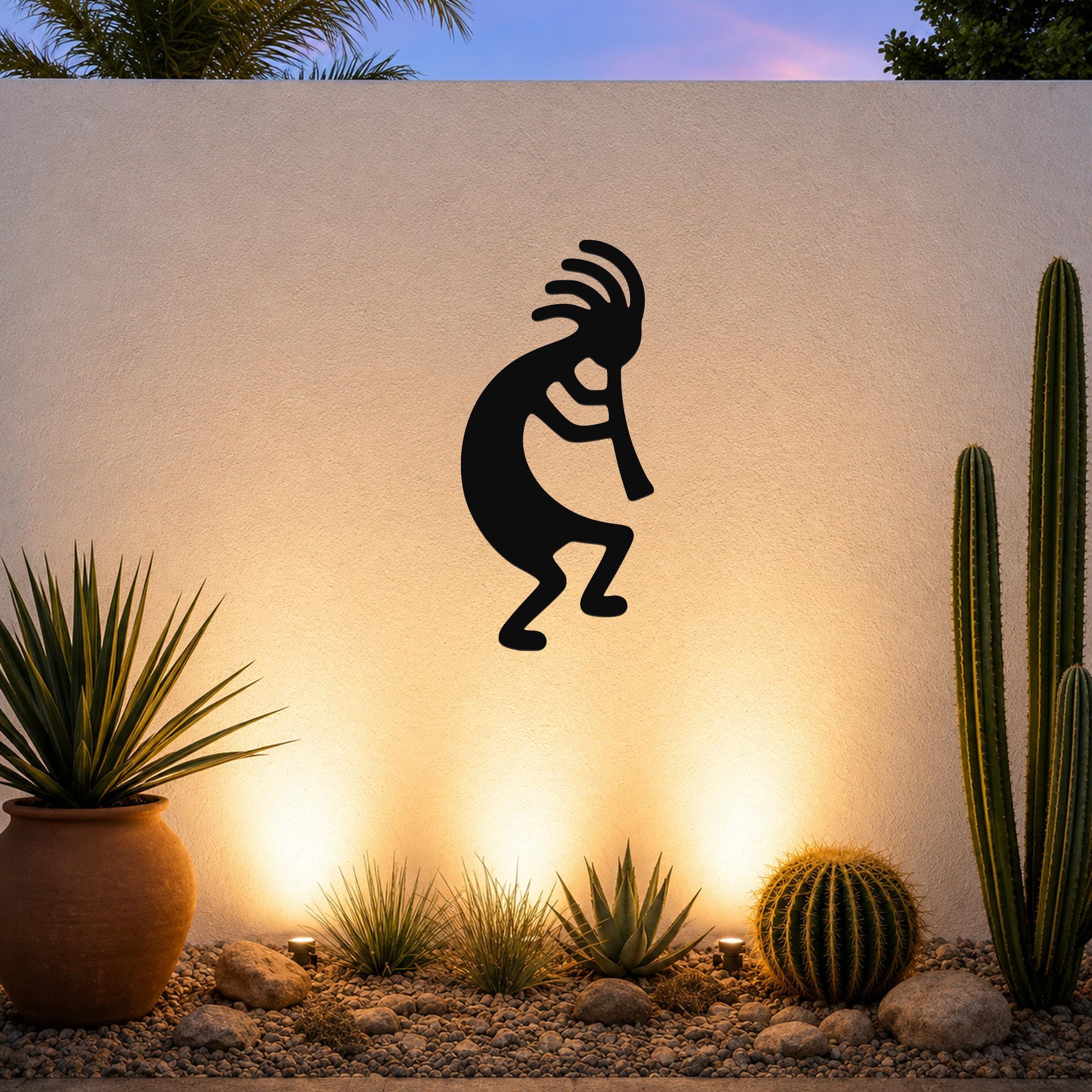 Black metal Kokopelli wall art silhouette with flute and feathered headdress, mounted on an outdoor wall above a desert garden with cacti and warm uplighting.