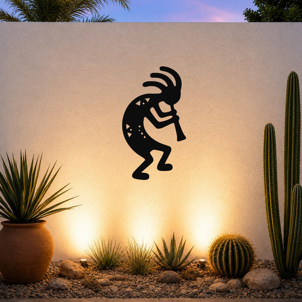 Black metal Kokopelli wall art silhouette with internal cutout details, mounted on a textured outdoor wall above a desert garden with cacti and warm uplighting.