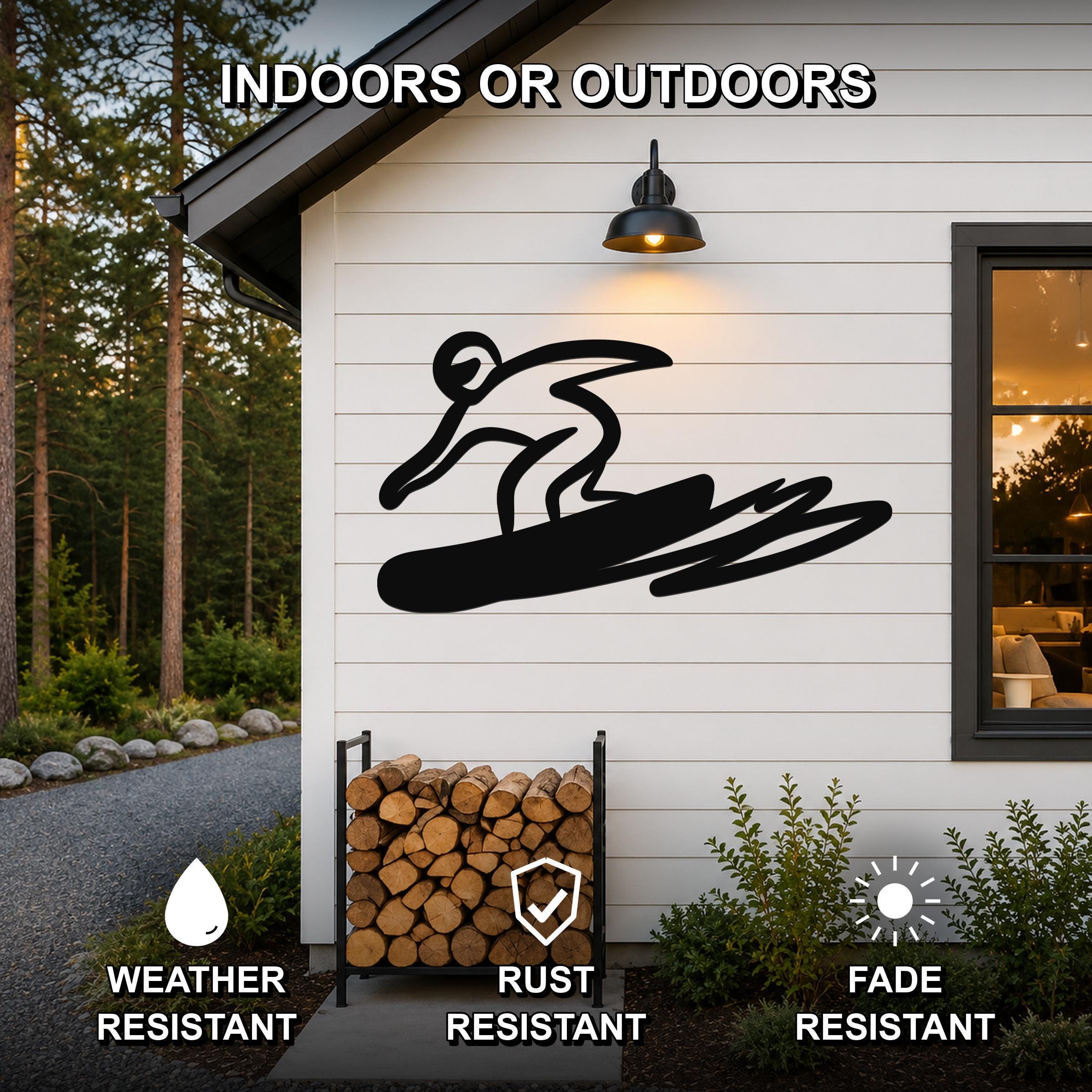 Black metal wall art featuring a bold silhouette of a snowboarder in motion, mounted on a white exterior wall above a firewood rack, with text indicating it is weather, rust, and fade resistant for indoor or outdoor use.