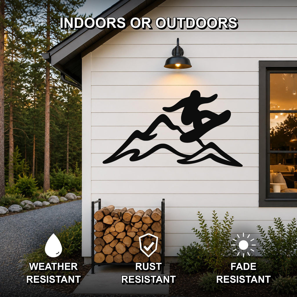 Black metal wall art featuring a snowboarder silhouette in motion over mountain peaks, mounted on a white exterior wall above a firewood rack, with text indicating it is weather, rust, and fade resistant for indoor or outdoor use.