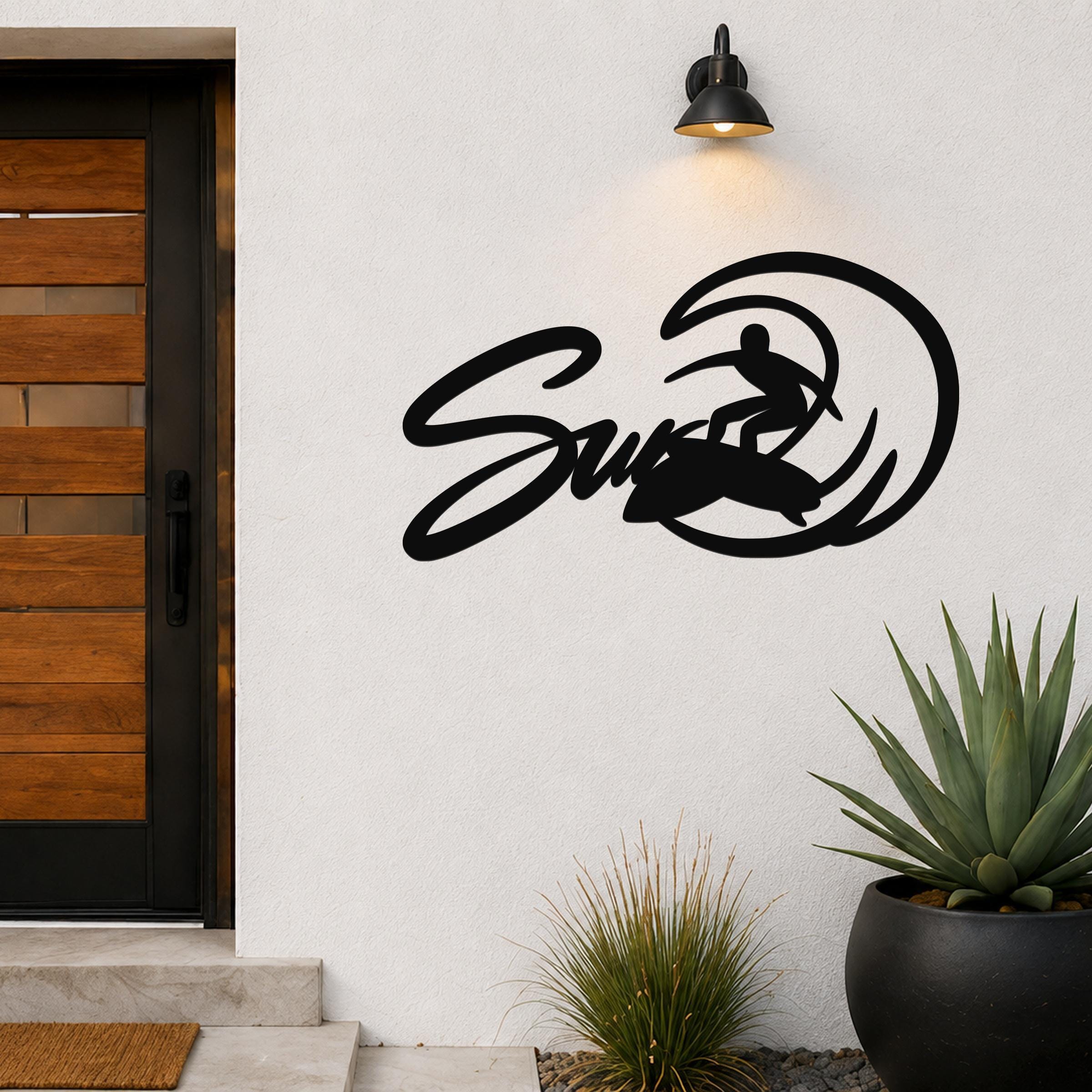 Black metal wall art featuring a surfer silhouette riding a wave, mounted on an outdoor wall beside a wooden door and potted plants.
