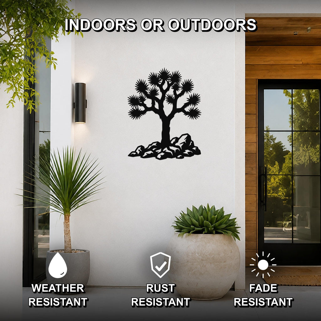 Black metal wall art featuring a Joshua tree silhouette with rocky base, mounted on a white exterior wall beside a glass door and potted plants, labeled as weather, rust, and fade resistant for indoor or outdoor use.
