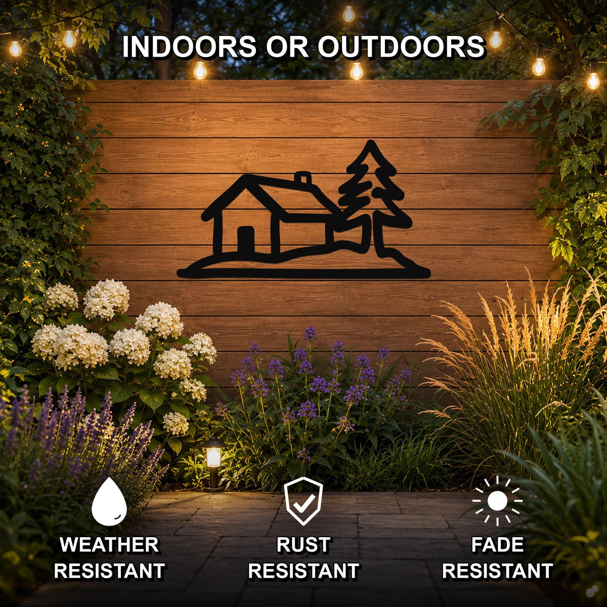 Black metal wall art silhouette of a cabin and tree, mounted on a wooden outdoor wall above a garden with flowers and string lights, weather resistant.
